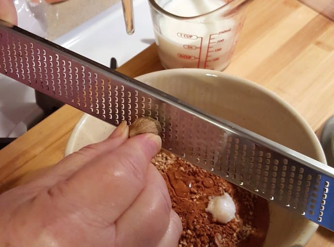 A hand with fresh nutmeg in it, using a Microplane to zest the nutmeg into a round beige bowl.