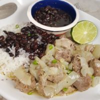 A white serving plate of Pressure Cooker Lechón Asado {Cuban Pork with Mojo} with rice, half a lime, and a bowl of black beans in a colorful round bowl.