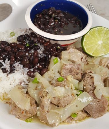 A white serving plate of Pressure Cooker Lechón Asado {Cuban Pork with Mojo} with rice, half a lime, and a bowl of black beans in a colorful round bowl.