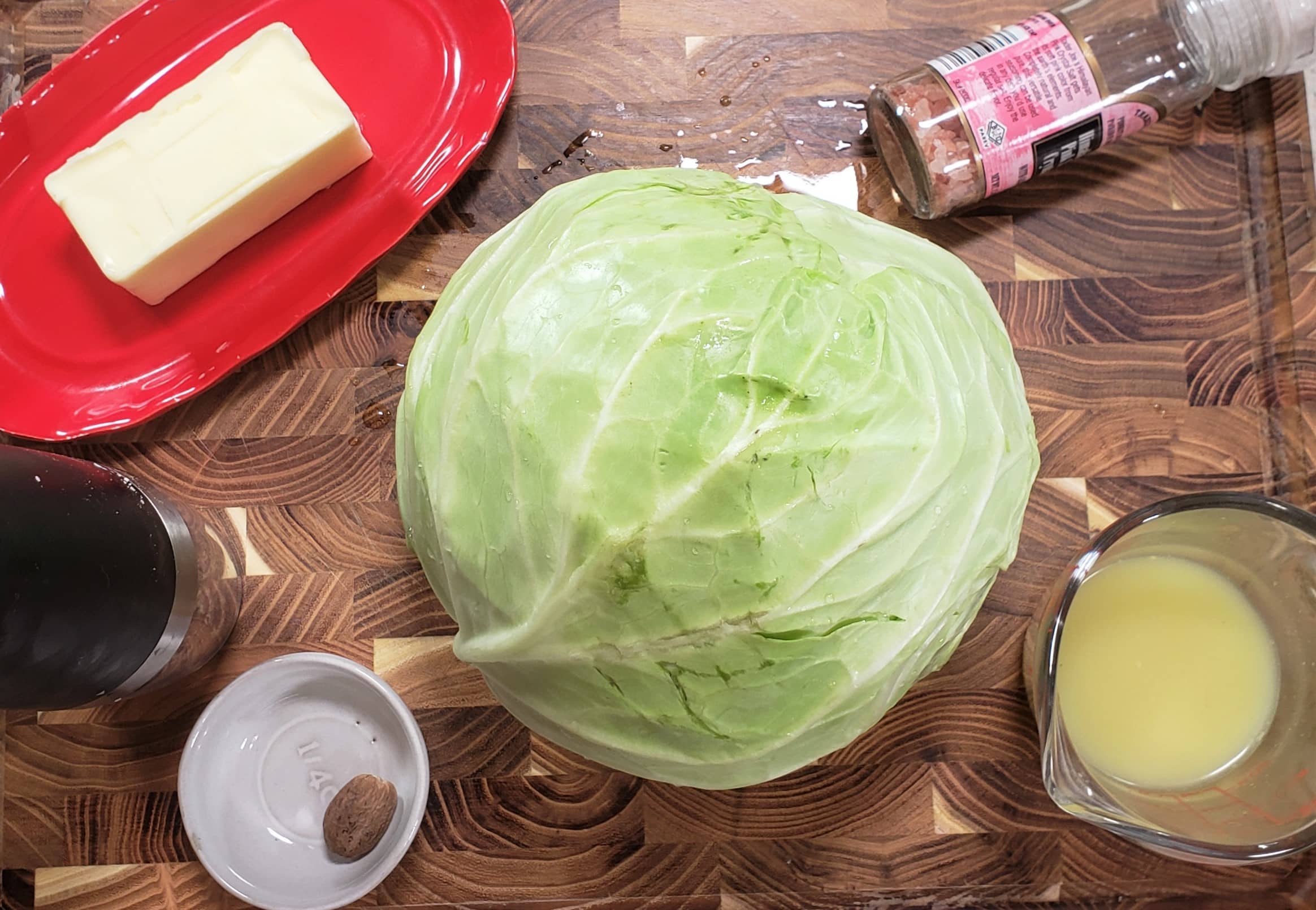 Ingredients for Instant Pot Buttered Cabbage on top of a wood cutting board. One head of cabbage, butter on a red butter dish, small white bowl with whole nutmeg, jar of Himalayan pink sea salt, glass measuring cup with chicken broth and black pepper grinder.