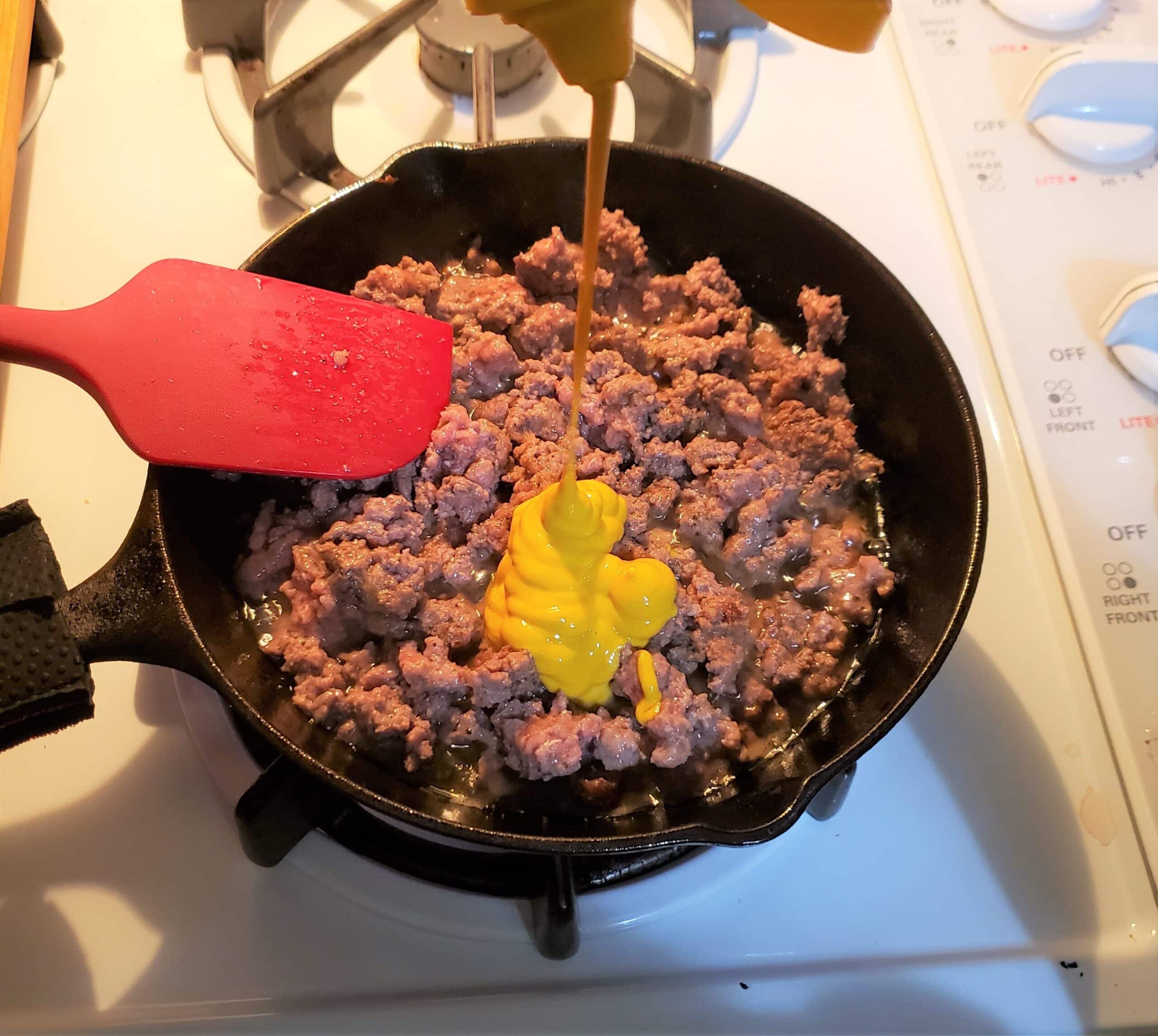 Yellow mustard being squirt into a cast ion pan filled with ground beef